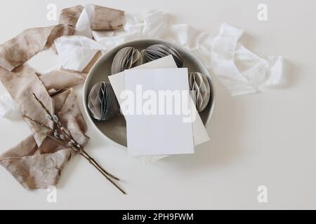 Frühjahrsgeschenk Stillleben. Ostergrußkarte, Einladungsmodell. Schüssel mit gefalteten Papierschmuck, Eier. Beigefarbenes Seidenband, Katzenfutter isoliert Stockfoto