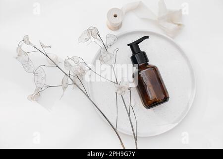 Braunglaskosmetikflasche. Shampoo-Pumpspender auf Keramikplatte. Trockene Lunariaflumen isoliert auf weißem Tischhintergrund. SPA-Bad natürlich Stockfoto