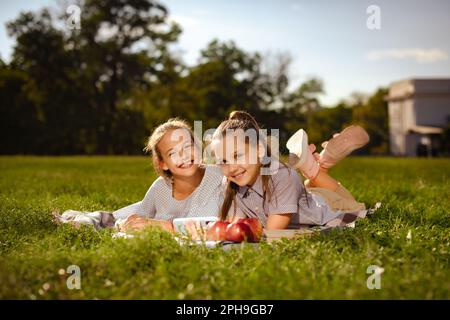 Ein paar Teenager-Freundinnen, die in die Kamera schauen und lächeln, haben Spaß auf grünem Gras im Park. Schulvlogger tragen lässige Hemden und ruhen sich aus Stockfoto