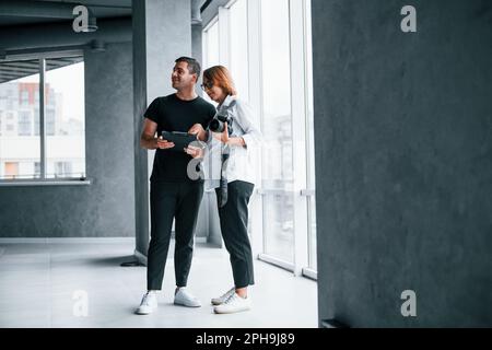 Frau in formeller Kleidung und mit Kamera in der Hand, die in einem leeren Raum mit einem Mann steht Stockfoto