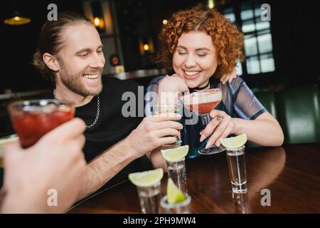 Lächelnde Freunde, die verschiedene Cocktails in der Nähe von Tequila-Shots mit Limette in der Bar, Stockbild Stockfoto