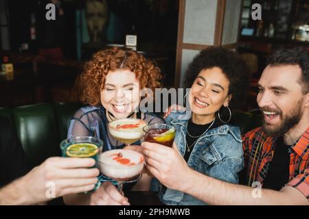 Positive interrassistische Freunde lächeln und stoßen mit verschiedenen Cocktails in der Bar an, Stockbild Stockfoto