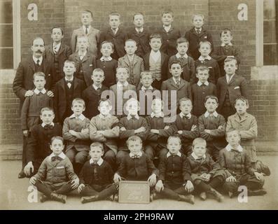 Colchester, Essex. Etwa 1910. Eine Klasse junger Schuljungen und ihr Lehrer posieren für ein Gruppenfoto vor ihrem Schulgebäude. Die Jungen besuchen die Magdalen Street Boys' School in Colchester, Essex. Ein Junge unterstützt eine Tafel mit dem Namen der Schule und der Klasse „Div 1“. Viele der Jungs tragen steife Leinenkragen und Norfolk-Jacken. Stockfoto