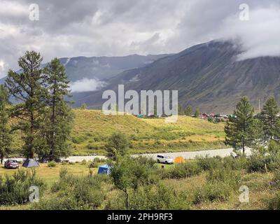 Orte zum Ausruhen mit Cottages und Zelten mit Autos von Touristen in der Nähe des Flusses in Altai nahe der Berge mit Wolken Stockfoto