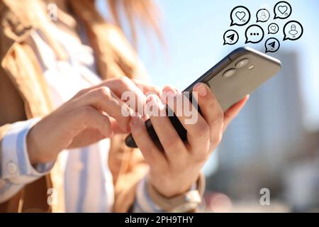 Verschiedene virtuelle Ikonen und junge Frau, die ihr Smartphone im Freien nutzt, Nahaufnahme. SMM-Konzept (Social Media Marketing) Stockfoto