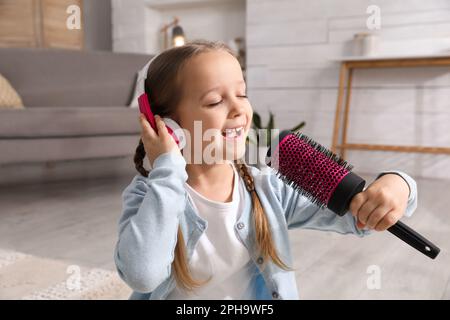 Süßes kleines Mädchen mit Kopfhörern und Haarbürste, die zu Hause singt Stockfoto