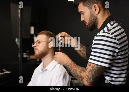 Friseur mit Bartöl in der Nähe des Kunden im Friseursalon. Professioneller Rasierservice Stockfoto