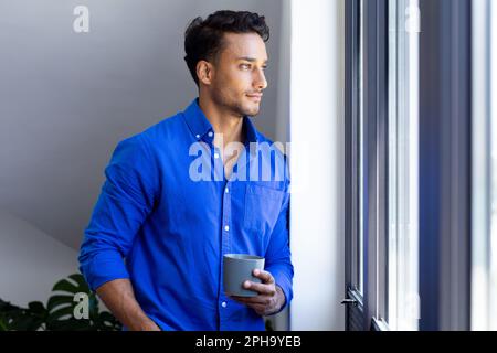 Ein glücklicher birassistischer Mann, der einen Becher hält und durch das Fenster schaut Stockfoto