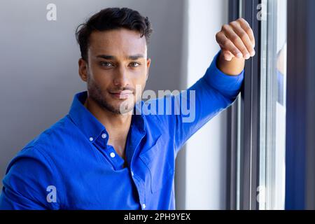 Porträt eines glücklichen birassischen Mannes, der sich ans Fenster lehnt und in die Kamera schaut Stockfoto