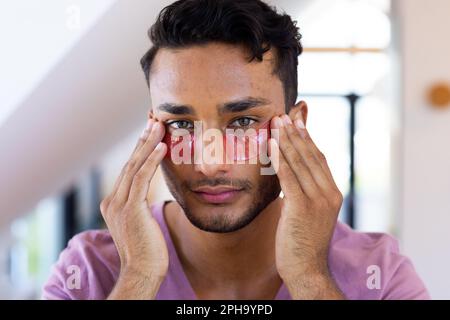 Glücklicher birassistischer Mann, der unter Augenmasken im Badezimmer aufträgt Stockfoto