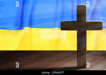 Bete für die Ukraine. Kreuz auf dem Holztisch gegen die ukrainische Flagge, Platz für Text Stockfoto