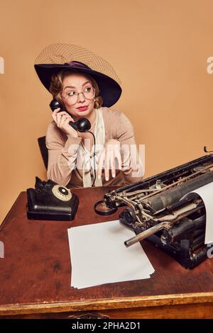Wunderschöne Frau in elegantem Vintage-Kostüm und Hut, die am Telefon spricht, mit einem unangenehmen Gesichtsausdruck auf beigefarbenem Hintergrund Stockfoto