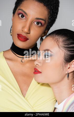 Porträt einer kurzhaarigen afroamerikanischen Frau, die in der Nähe eines asiatischen Freundes in die Kamera schaut, mit roten Lippen isoliert auf grauem Stockbild Stockfoto