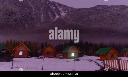 Altai, Russland - 02. Oktober 2022 Auto honda hrv steht neben Ferienhäusern mit einem leuchtenden Fenster in der Nähe des Waldes unter einem Berg mit Nebel und Schnee Stockfoto