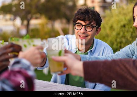 Ein fröhlicher junger Mann mit Brille lächelt an einer Bar im Freien, mit den Händen seiner Freunde und Drinks im Hintergrund - und genießt ein geselliges Treffen in einem Stockfoto