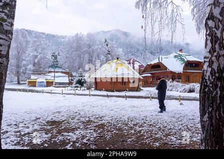 Eine ältere Frau läuft zwischen den Birken des runden Hauses der Nagel vor dem Hintergrund von Holzhäuschen im Schnee im Winter im Altai in Stockfoto