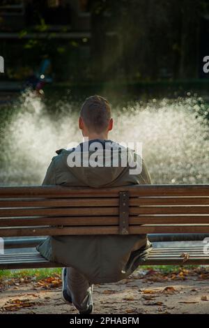 Ein Mann auf der Bank im Park, der auf den Brunnen blickt Stockfoto