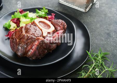 Steaks, Osso Buco. Rinderscheiben, gegrillter Osso Buco oder Porterhouse-Fleischsteak mit garniertem Salat und pommes frites auf schwarzem Marmorbrett auf altem Holz Stockfoto