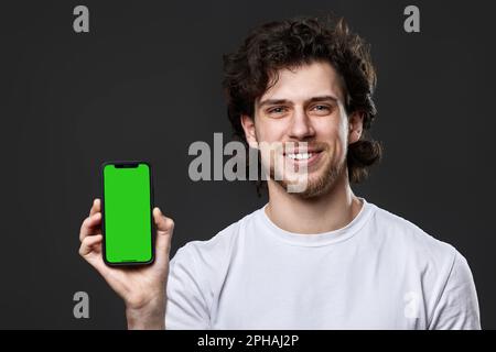 Glücklicher junger Mann, der ein Telefon mit grünem Bildschirm auf dunkelgrauem Hintergrund zeigt Stockfoto