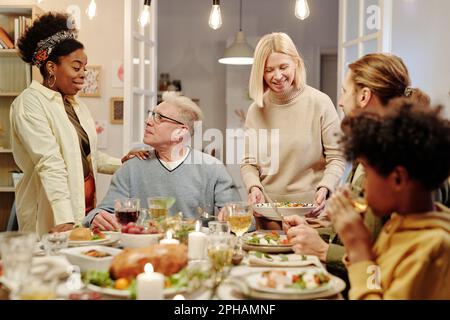 Junge schwarze Frau, die Hand an der Schulter eines erwachsenen Mannes hält und mit ihm spricht, während sie am Esstisch steht und mit hausgemachtem Essen serviert wird Stockfoto