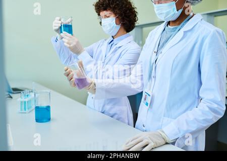 Zwei junge interkulturelle Klinikinnen in Labormänteln und Schutzmasken, die mit Flüssigkeiten in Reagenzgläsern arbeiten, während sie am Arbeitsplatz stehen Stockfoto