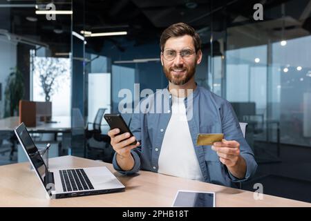 Ein lächelnder junger Mann hält ein Telefon und eine Kreditkarte. Steht im Büro, wählt am Telefon, tätigt Online-Käufe, bestellt, prüft das Konto Stockfoto