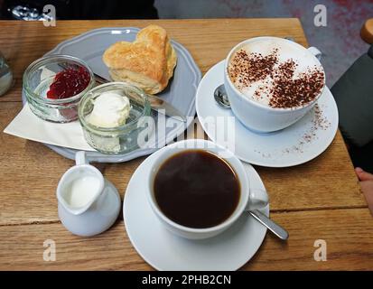 Nahaufnahme Scone mit Erdbeermarmelade und Clotted Curd Cream serviert mit heißem schwarzen Kaffee und Latte Stockfoto