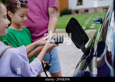 Nahaufnahme eines Vaters, der das Netzkabel mit seinen kleinen Töchtern hält und sein Elektroauto lädt. Stockfoto