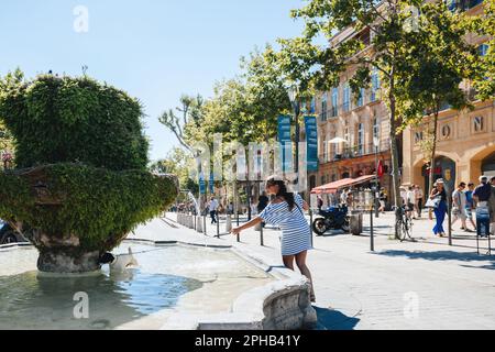 Aix-en-Provence, Frankreich - 17. Juli 2014: Die Menschen kommen gemütlich in der heißen Sonne des Cours Mirabeau, Aix-en-Provence vorbei. Bäume und der Brunnen des Neufs Kanonen zieren einen einzigartigen platz in der Stadt, umgeben von atemberaubender Architektur. Stockfoto