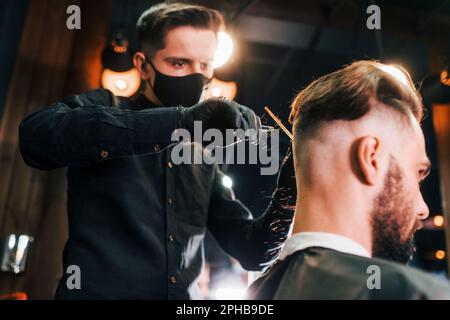 Ein junger bärtiger Mann sitzt im Friseursalon und lässt sich die Haare schneiden, von einem Kerl mit schwarzer Schutzmaske Stockfoto