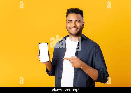 Fröhlicher, aufgeregter junger indischer Mann, der auf die Kamera schaut, mit dem Finger auf den leeren Telefonbildschirm zeigt, auf Werbeschriften achtet, Werbung für neue Werbeaktionen macht, Verkaufsangebote präsentiert, isoliert auf Gelb steht Stockfoto