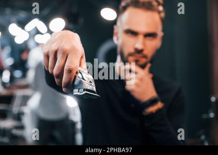Ein junger bärtiger Mann, der im Friseursalon steht und den Haarschneider hält Stockfoto