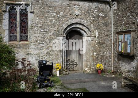 Am 19. März 2023 in Manaccan, Cornwall, England, wurde ein Mobilitätsroller vor der Tür der anglikanischen Kirche St. Manaccus & St. Dunstan im Cornish Village Manaccan abgestellt. Stockfoto