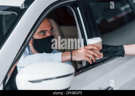 Ich kaufe ein frisches Getränk. Junger gutaussehender Mann in formeller Kleidung und Schutzmaske sitzt in einem brandneuen Automobil Stockfoto