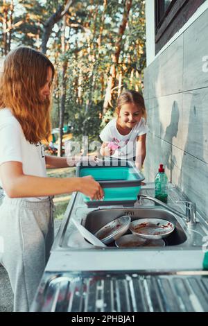 Teenager-Mädchen waschen das Geschirr Töpfe und Teller mit Hilfe ihrer jüngeren Schwester in der Outdoor-Küche während des Urlaubs auf Camping. Lagerleben. Schwester Stockfoto