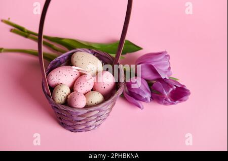 Stillleben. Osterhintergrund mit rosa Pastelltönen und beigen Wachteleiern in einem lila Korb mit einem Strauß Tulpen Stockfoto