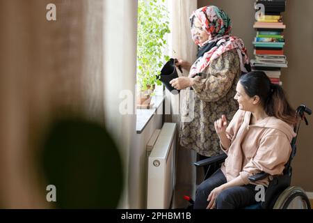Eine Frau mit Querschnittslähmung, die zu Hause mit der Mutter spricht, die die Pflanze tränkt Stockfoto