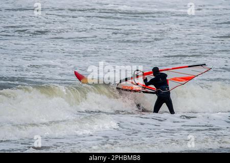 Windsurfer in schwarzem Neoprenanzug, der bei Wind und Wintersturm an der Nordseeküste klassisches Windsurfen praktiziert Stockfoto