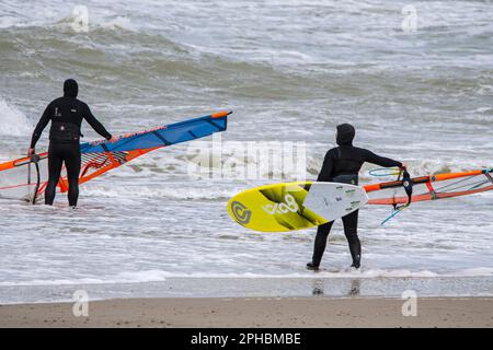 Zwei Windsurfer in schwarzen Neoprenanzügen betreten das Wasser, um klassisches Windsurfen an der Nordseeküste bei Wind und Wintersturm zu üben Stockfoto