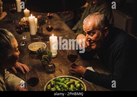 Blick aus dem Blickwinkel auf einen erwachsenen Mann, der mit einer älteren Freundin spricht, die während der Party am Esstisch sitzt Stockfoto