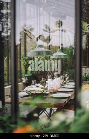 Tischaufstellung für Dinnerparty im Hinterhof-Gewächshaus, durch Glasfenster gesehen Stockfoto