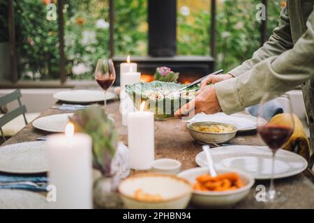 Hände eines Seniorenmannes, der eine Salatschüssel hält, während er den Esstisch für die Party vorbereitet Stockfoto