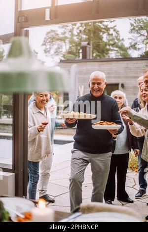 Ein glücklicher, reifer Mann, der Essteller mit sich führt und mit Freunden während der Dinnerparty spaziert Stockfoto