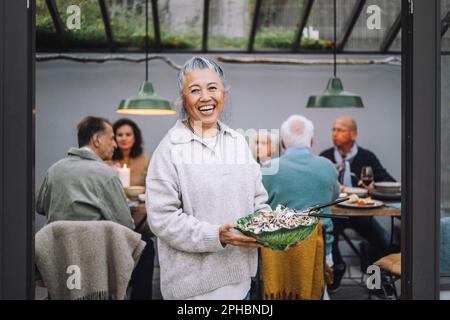 Porträt einer fröhlichen Seniorin, die Salatschüssel auf der Dinnerparty hält Stockfoto