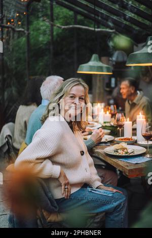 Seitliches Porträt einer lächelnden Seniorin, die während der Dinnerparty auf einem Stuhl sitzt Stockfoto