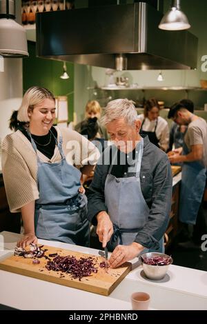 Lächelnde nicht-binäre Person, die während des Kochkurses in der Küche auf einen männlichen Schüler schaut, der Gemüse hackt Stockfoto