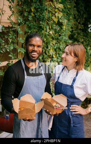 Lächelnde Chefköchin mit Hand in der Hüfte, die den männlichen Besitzer ansieht, der im Restaurant Lebensmittelboxen hält Stockfoto
