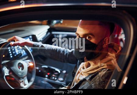 In schwarzer Schutzmaske. Ein gut aussehender, unrasierter Mann in modischer Kleidung, der sein Auto fährt Stockfoto