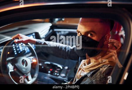 In schwarzer Schutzmaske. Ein gut aussehender, unrasierter Mann in modischer Kleidung, der sein Auto fährt Stockfoto