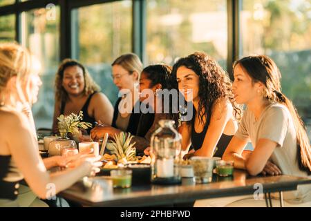 Fröhliche, multirassische weibliche Freunde sitzen am Esstisch und frühstücken im Rückzugszentrum Stockfoto